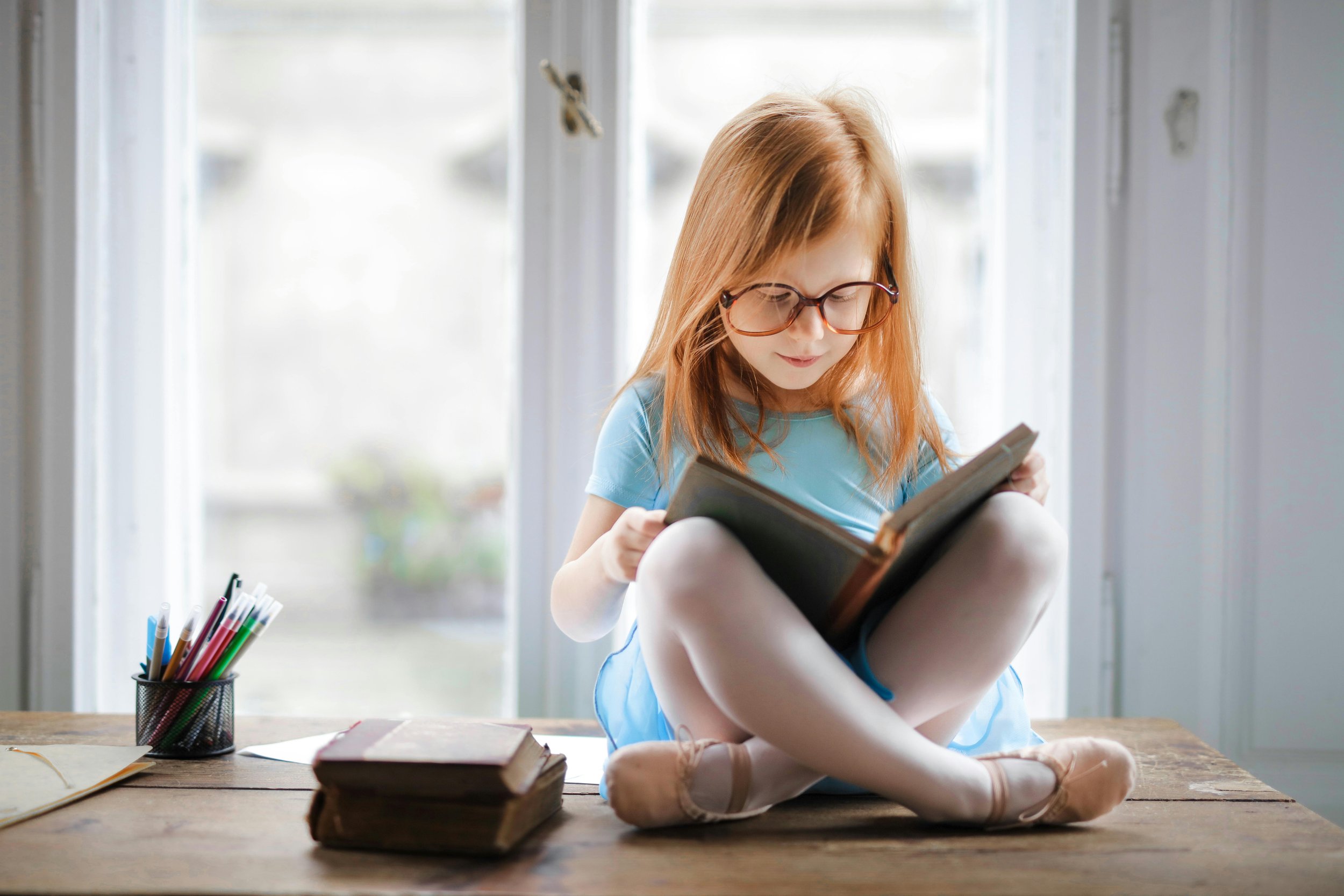 A young girl with red hair and glasses wearing a blue dress sitting cross-legged on a wooden table, reading a book.