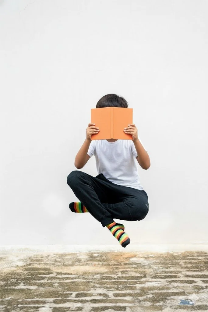 A child sitting cross-legged on the floor, holding an orange book up to their face, with only dark hair visible, against a plain white wall.