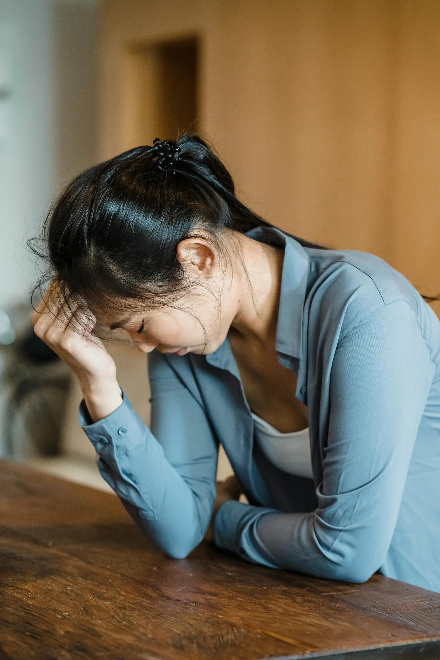 A woman sitting at a wooden table with her head resting on her hand, appearing distressed or upset.