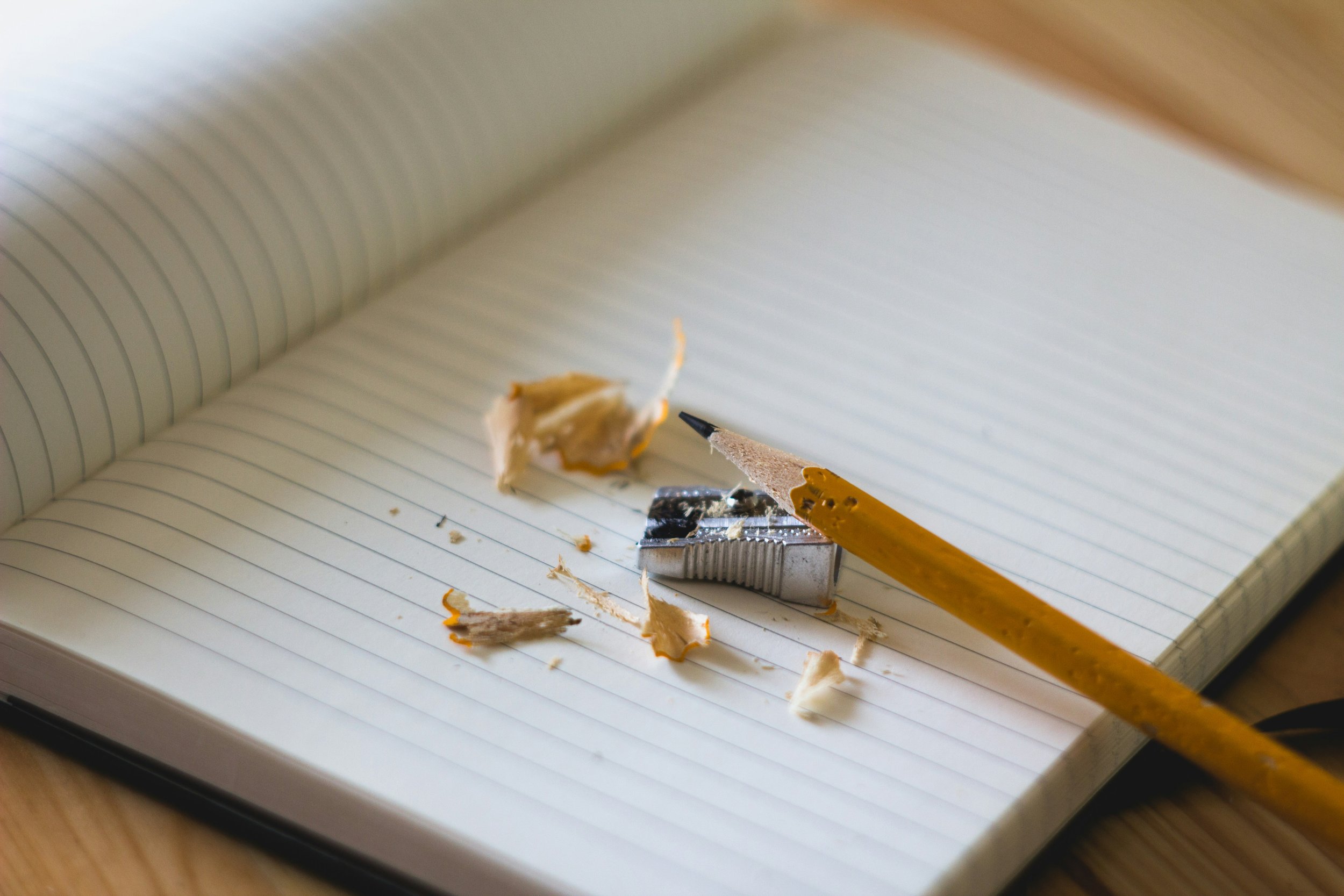 Open notebook with lined pages, surrounded by pencil shavings, a yellow pencil, and a metal sharpener on a wooden surface.