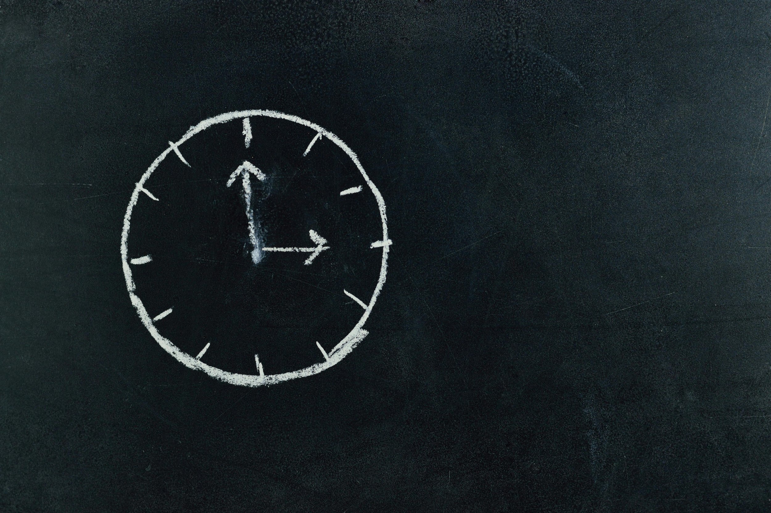 A clock drawn with white chalk on a black chalkboard, showing the time as 1:00.