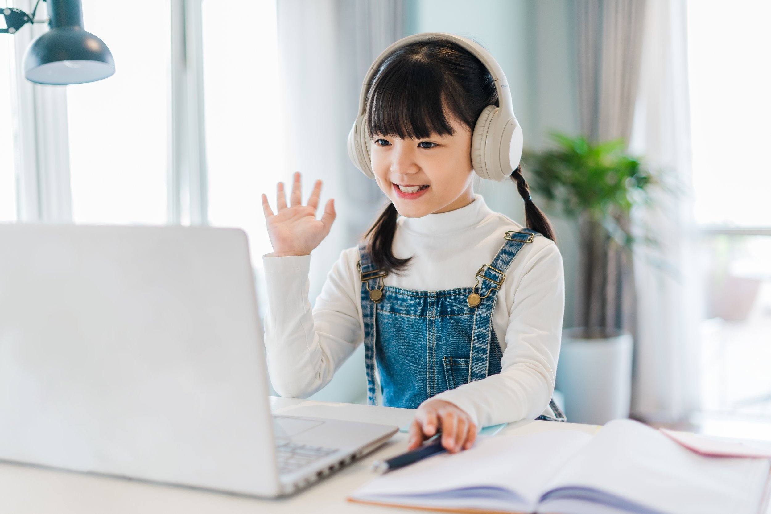 Young girl with long dark hair in pigtails wearing a white turtleneck and denim overalls, using a laptop, wearing large beige headphones, and waving at the screen, sitting at a desk in a bright room with a potted plant and curtains in the background.
