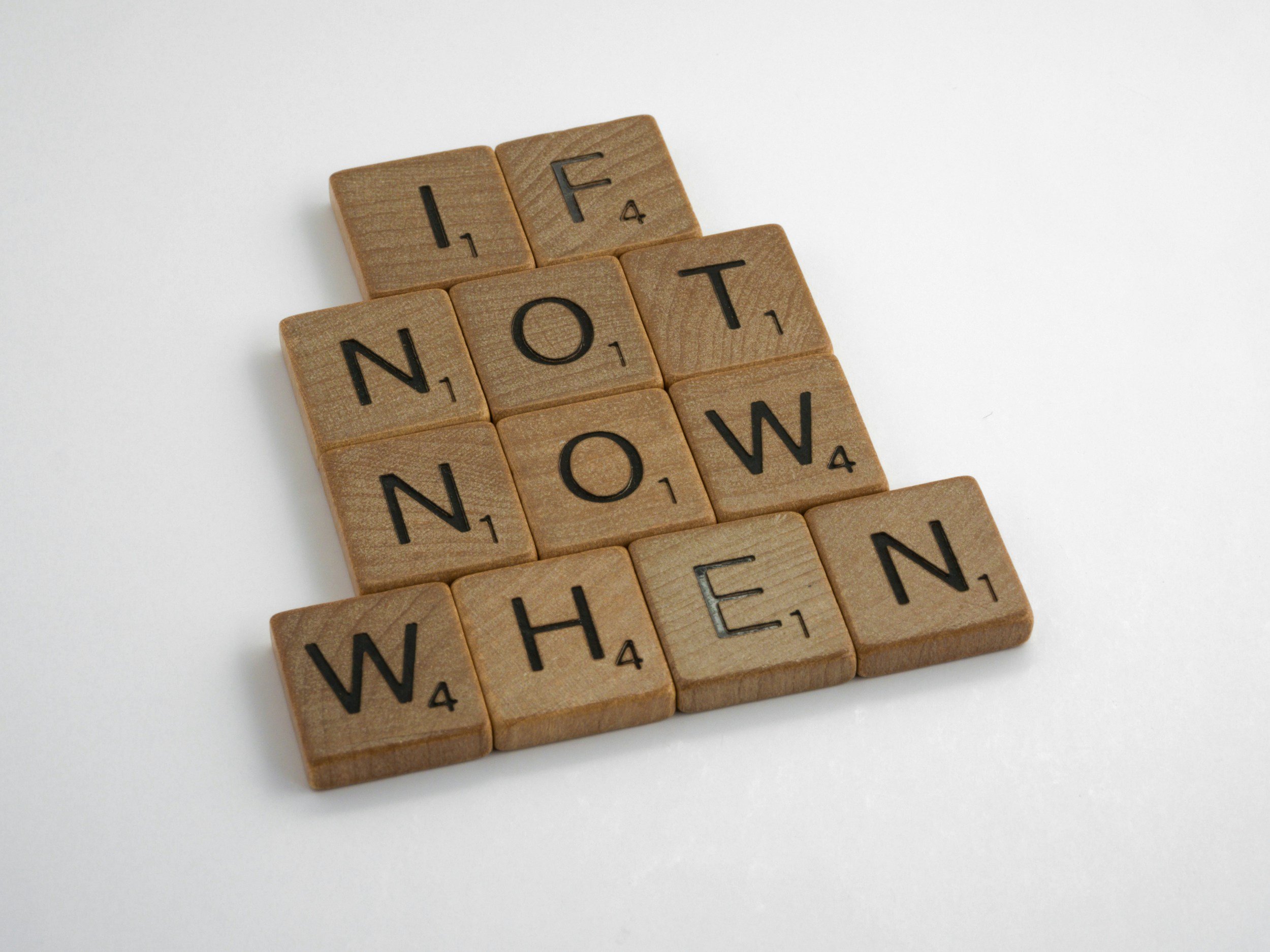 Wooden Scrabble tiles spelling out 'I F T NOT W H EN' on a white surface.
