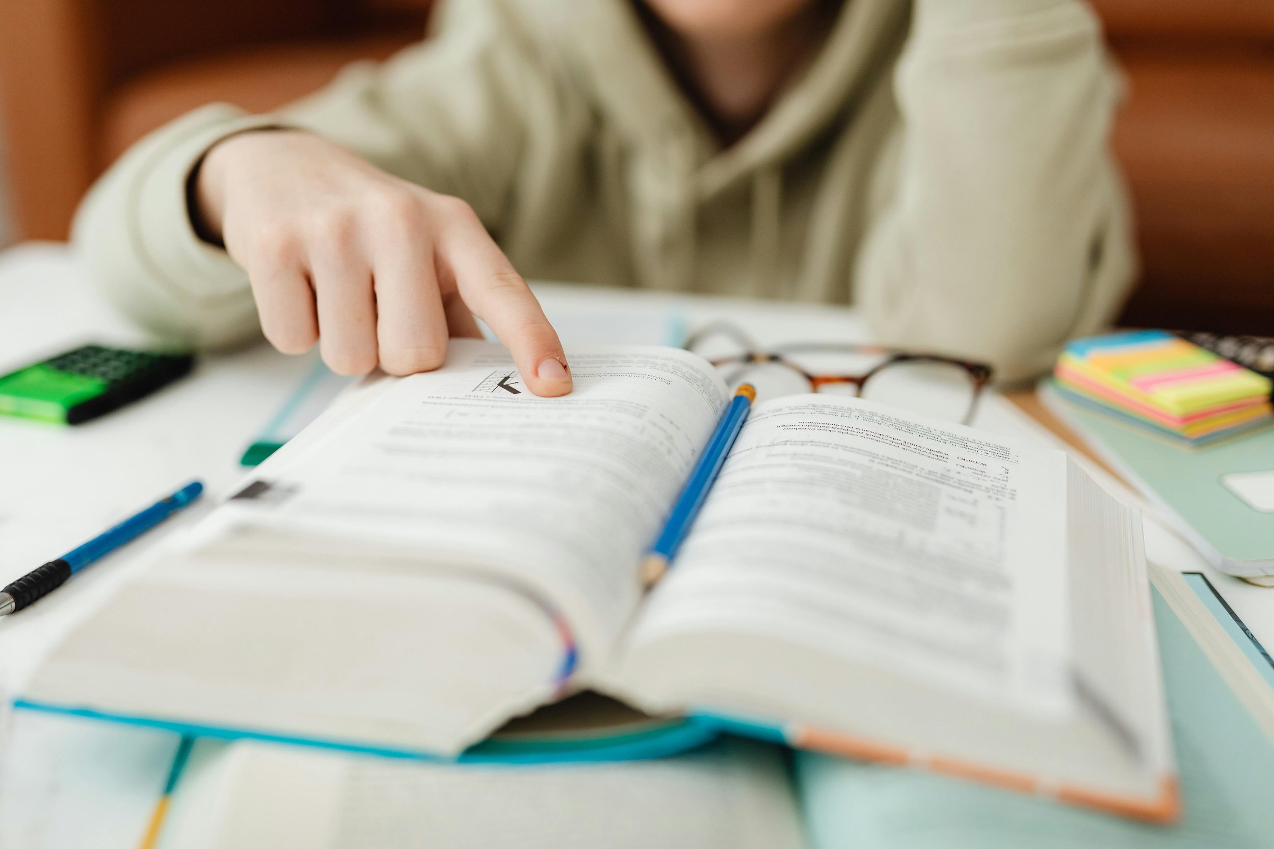 A person studying with an open book, a blue pencil resting on the page, and various stationery items on the desk, including a calculator, sticky notes, and a pair of glasses.