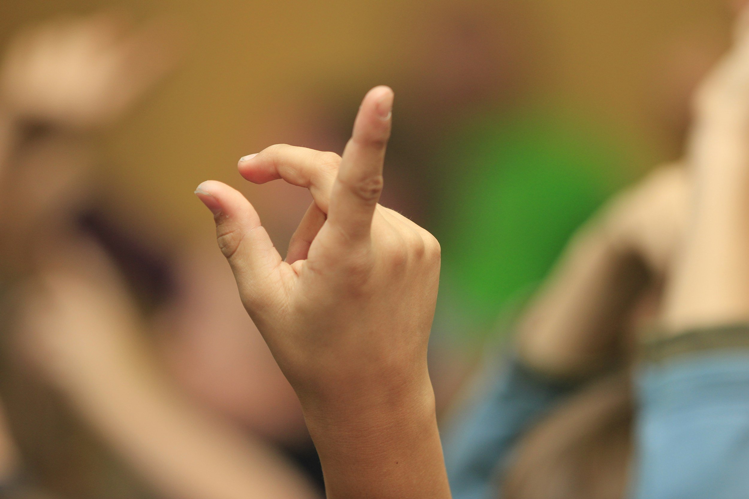 Close-up of a person's hand making an 'OK' gesture with the thumb and index finger touching, with blurred people in the background.